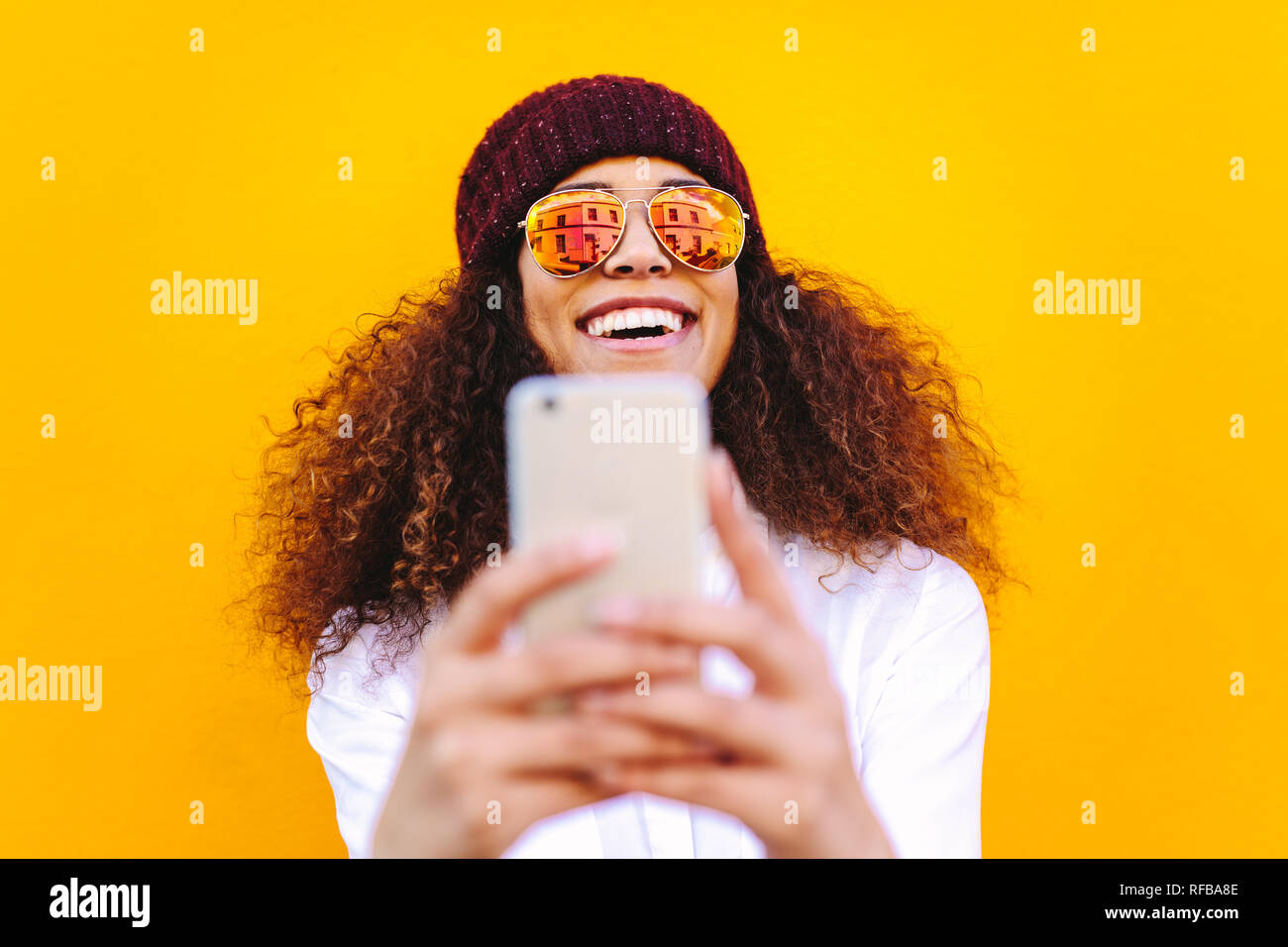 Stilvolle afrikanische Frau mit Handy zu nehmen selfie auf gelbe Wand. Attraktive Frau mit Hut und Sonnenbrille vor gelbem Hintergrund ein Stockfoto