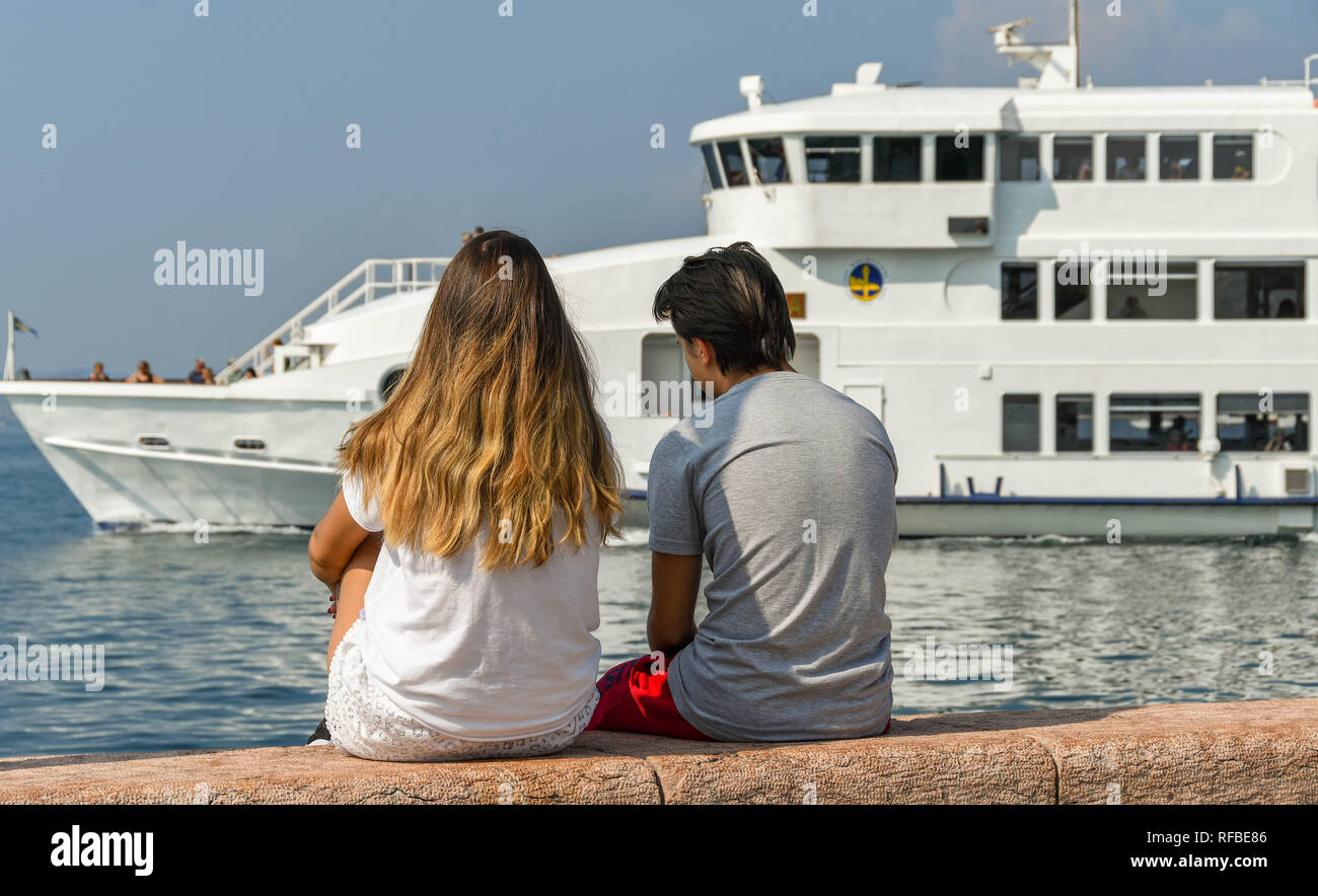 Gardasee, Italien - September 2018: Zwei junge Leute zusammen sitzen auf der Hafenmauer in der Stadt Garda am Gardasee. Im Hintergrund, ein Pass Stockfoto