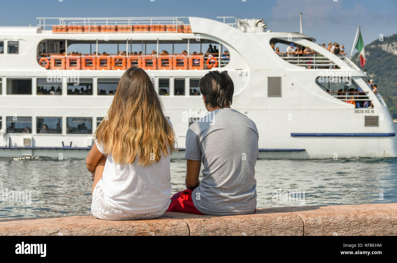 Gardasee, Italien - September 2018: Zwei junge Leute zusammen sitzen auf der Hafenmauer in der Stadt Garda am Gardasee. Im Hintergrund, ein Pass Stockfoto