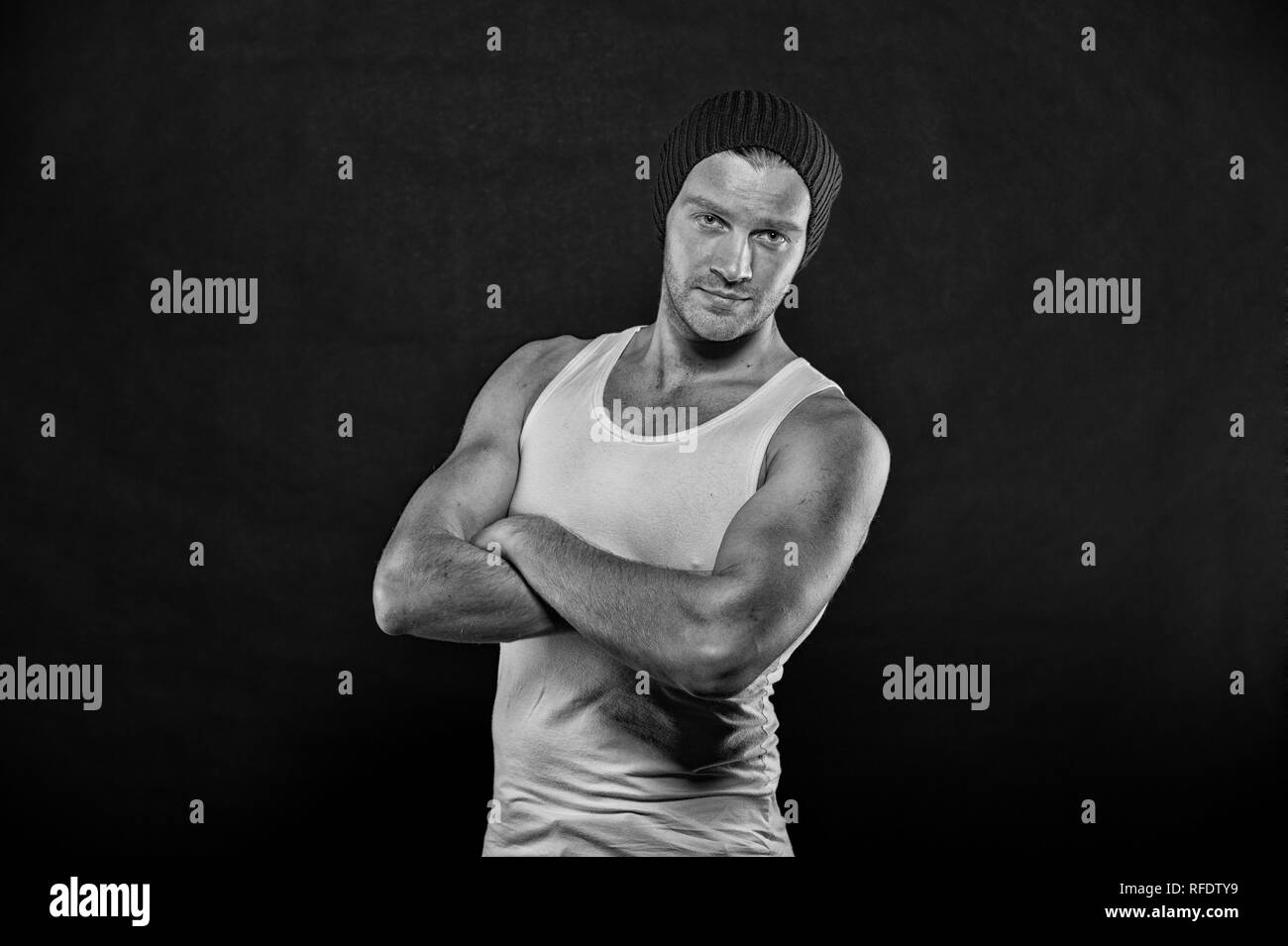 Macho stand mit starken Armen gefaltet. Mann in trendige Weste und Hut auf dunklem Hintergrund. Sportler mit Borsten auf ernstes Gesicht. Sport und Fitness. Mode Stil und Trend, Schwarz und Weiß. Stockfoto