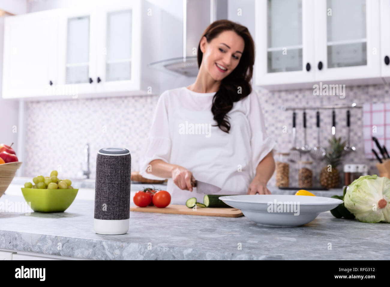 Glückliche Frau im Gespräch mit Voice Assistant beim Schneiden Gurken mit Messer Stockfoto