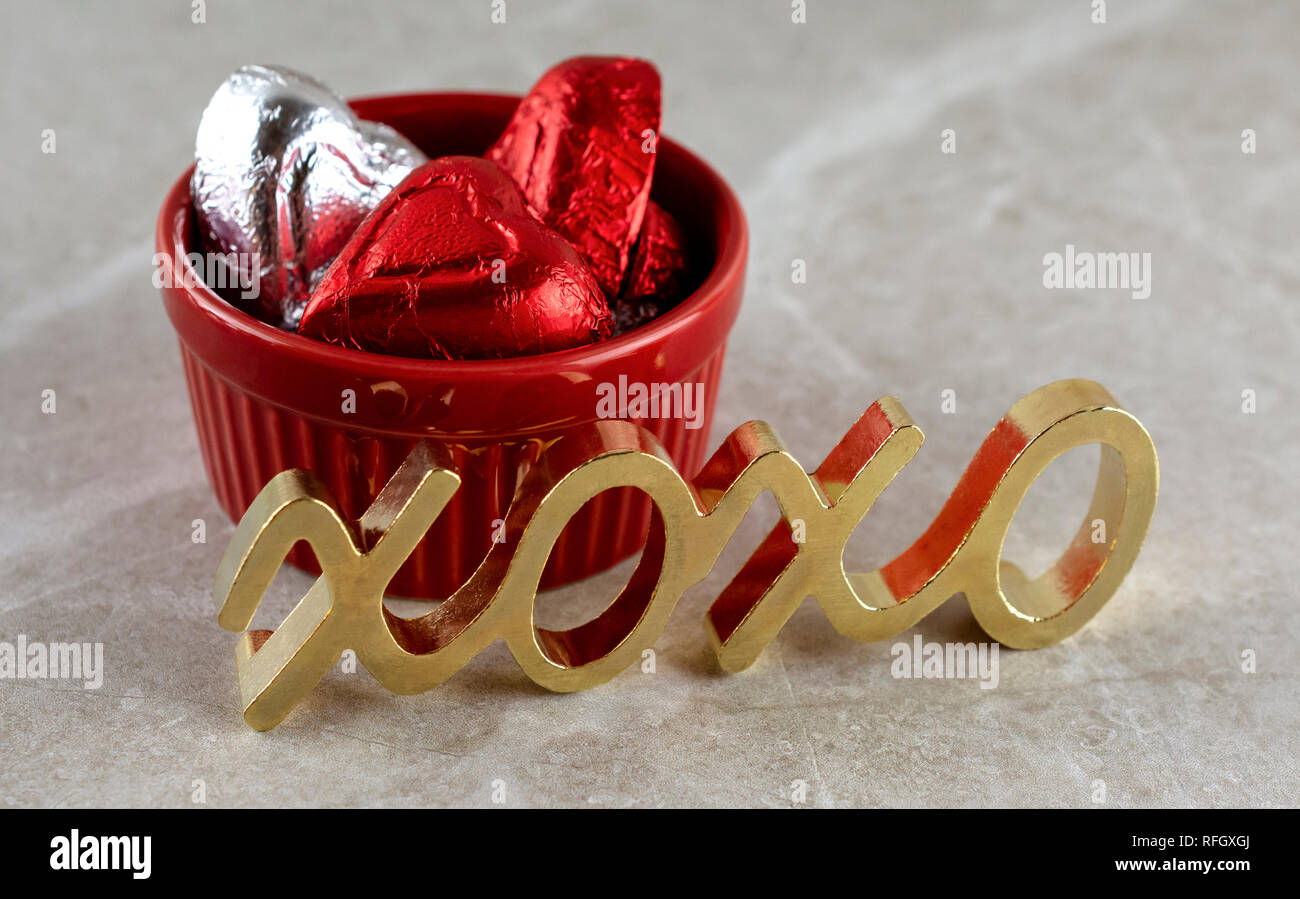 Nahaufnahme von roten ramekin mit Rot und Silber herzförmige Pralinen gefüllt. Gold Metall Symbol für Umarmungen und Küsse im Vordergrund. Tan Hintergrund Stockfoto