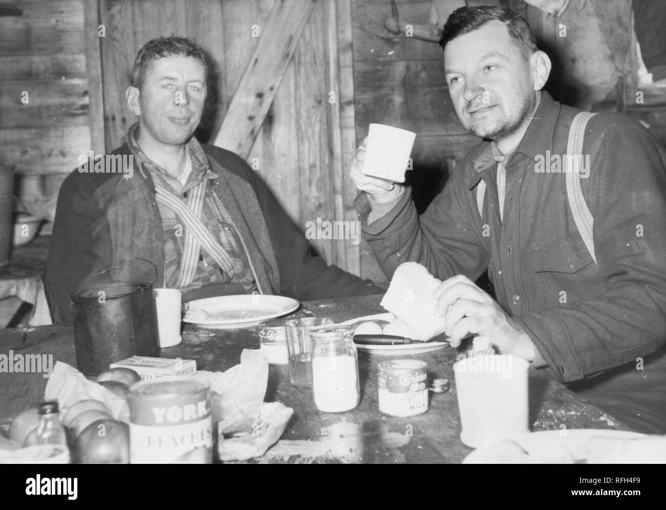 Schwarz-weiß Foto von zwei unrasiert, Männer im mittleren Alter, jedem tragen wollene Shirts und Hosenträgern, sitzen gemeinsam in einer Blockhütte oder Shack, essen eine Mahlzeit in einem unordentlichen Tabelle, mit verschüttetem Zutaten, ein Glas Milch oder Zucker, und eine Dose "Marke Pfirsiche York' im Vordergrund sichtbar, während einer Jagd und Angeln in Alaska, USA, 1955 entfernt fotografiert. () Stockfoto