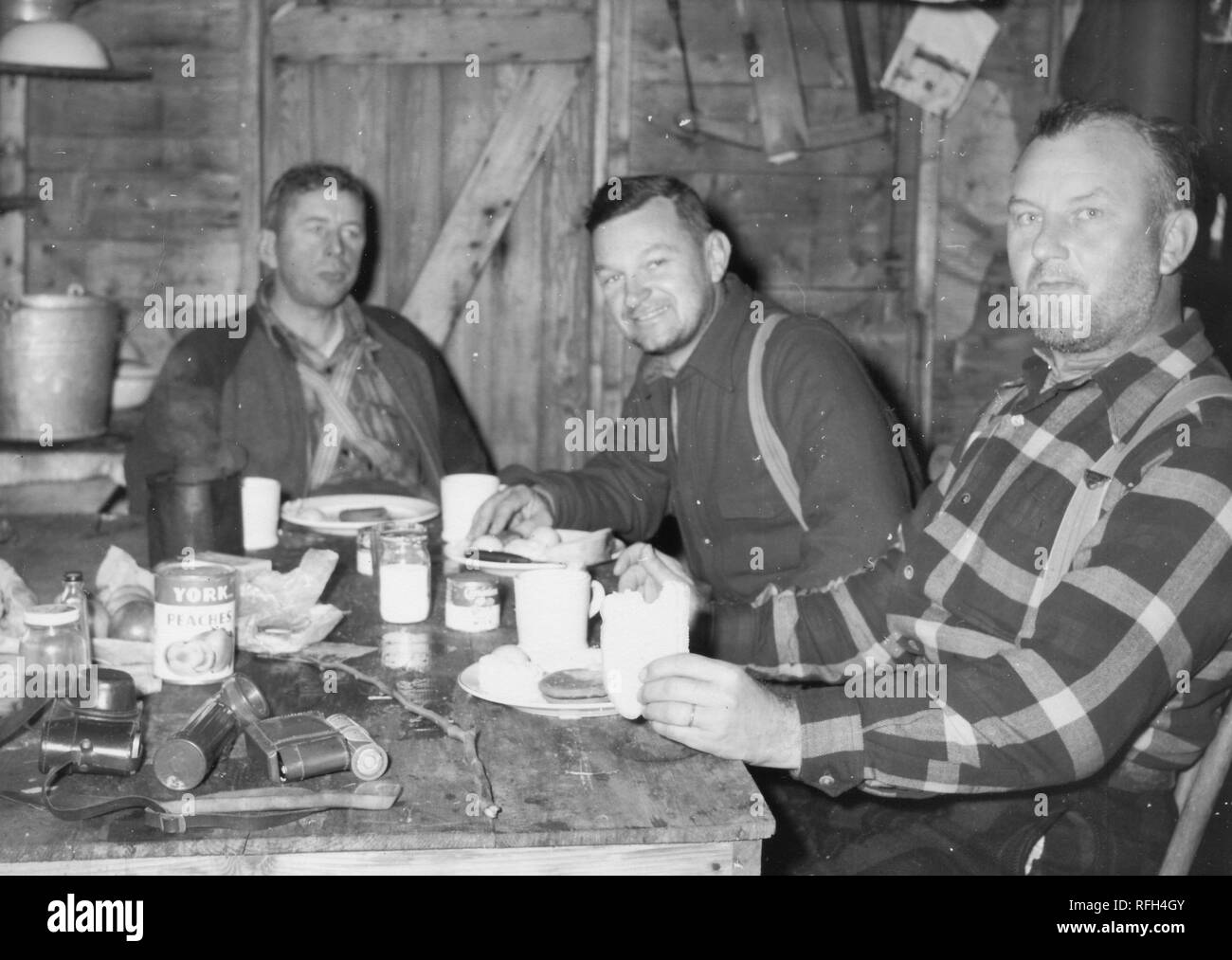 Schwarz-weiß Foto von drei unrasiert, Männer mittleren Alters (einer in der Mitte lächelnd) Jedem tragen wollene Shirts und Hosenträgern, sitzen gemeinsam in einer Kabine oder Shack eine Mahlzeit zu essen, mit der Kamera ein Stick, und eine Dose "Marke Pfirsiche York" auf dem Tisch vor ihnen, während einer Jagd und Angeln in Alaska, USA, 1955 entfernt fotografiert. () Stockfoto