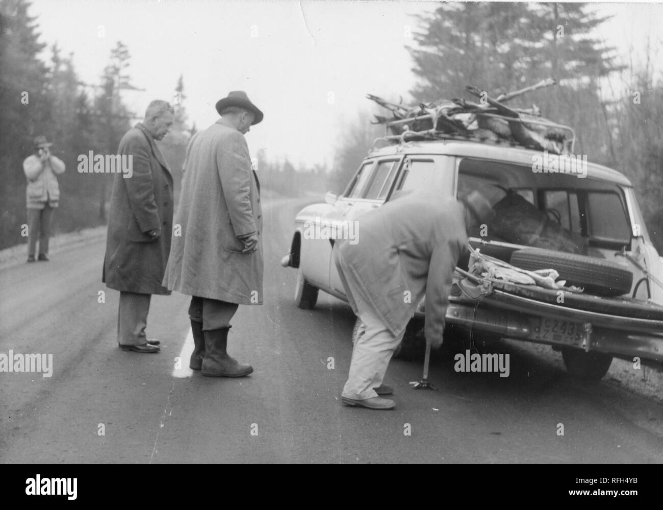 Schwarz-weiß Foto von vier Männern, jeder trägt einen Mantel oder Jacke,  die auf einem leeren Strecke von der Straße mit einem geparkten Kombi, die  über die Körper von mehreren Hirsch in seiner