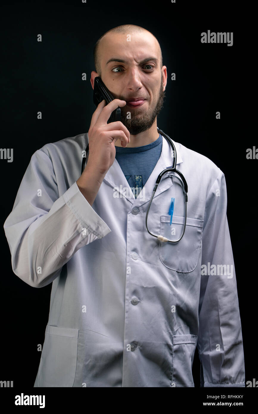 Crazy Arzt zeigt lustige Grimasse während der Verwendung des Telefons und Stethoskop angezeigt. Junge Praktiker in Uniform Stockfoto