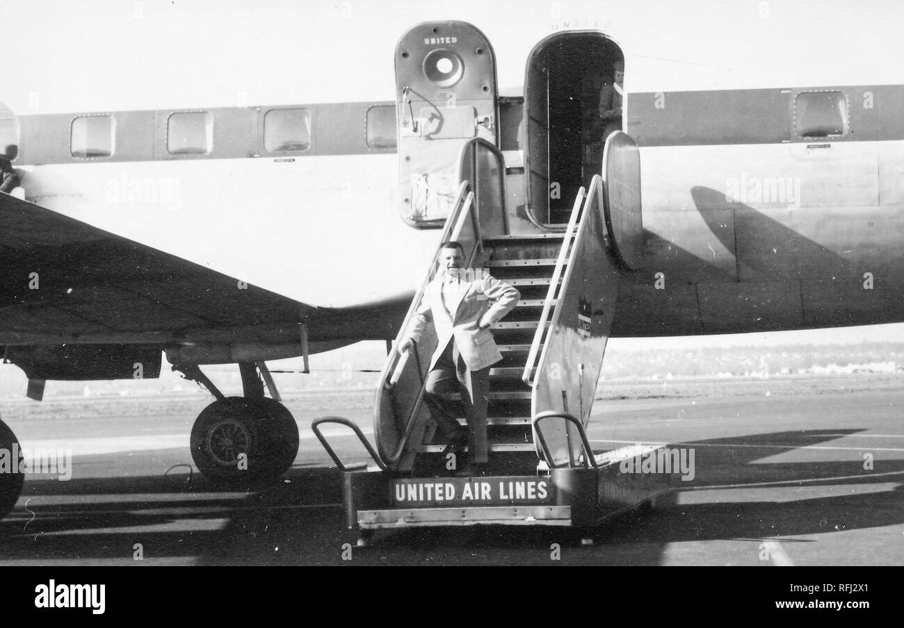 Schwarz-weiß Foto eines Dunkelhaarige, Mann mittleren Alters, in voller Länge gesehen, Anzug, stehend in contrapposto mit einer Hand auf seiner Hüfte darstellen, auf der untersten Stufe der ein Flug der Flugzeug Treppen oder landesteg, mit einem Mitglied der Kabinenbesatzung teilweise im offenen Flugzeug Tür sichtbar im Hintergrund, und das Logo "United Air Lines' auf der niedrigsten Riser unter der man Füße sichtbar, während einer Jagd und Angeln in Alaska, 1955 entfernt fotografiert. () Stockfoto