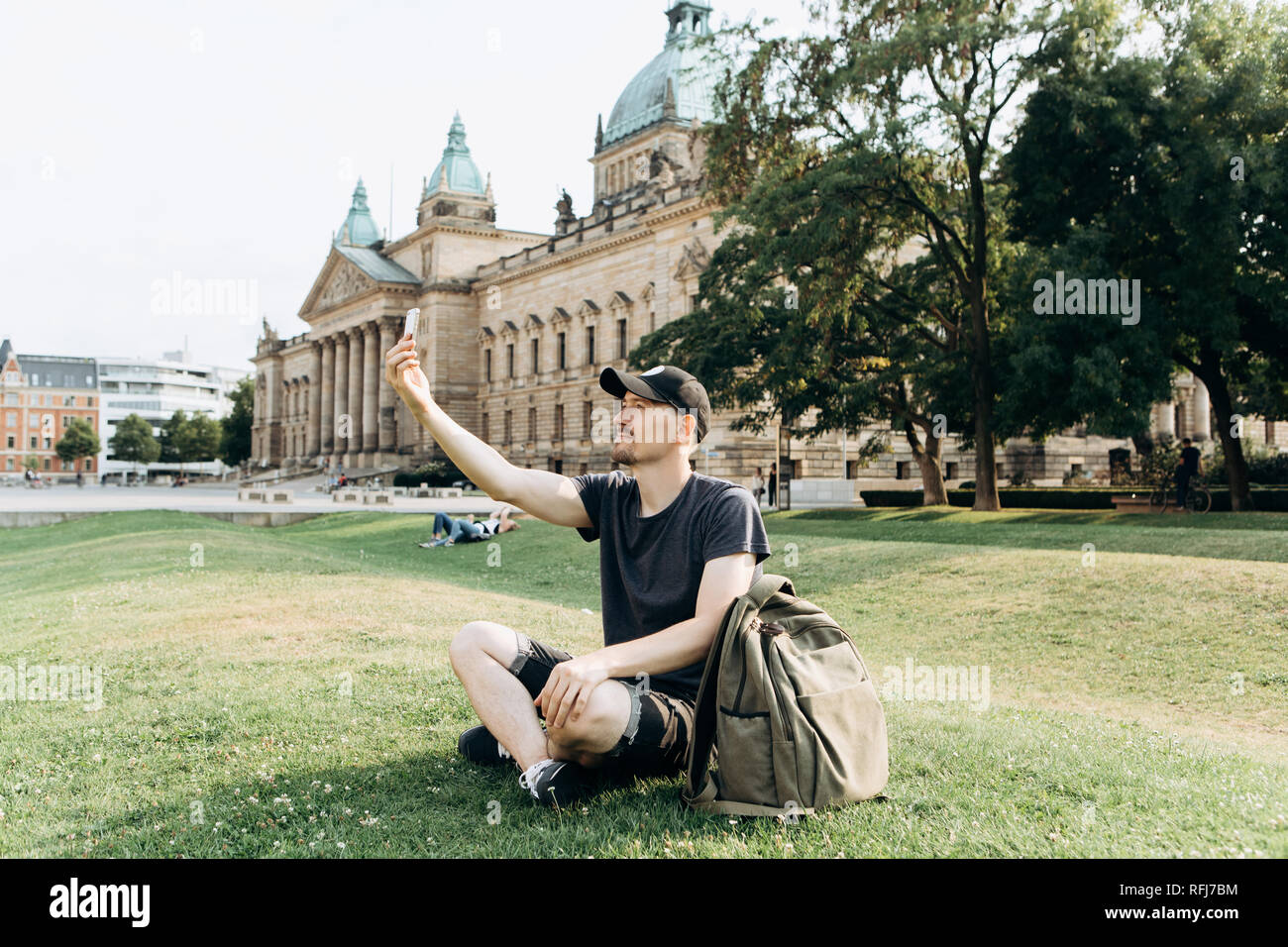 Eine junge männliche Touristen oder Studenten sitzt auf dem Gras außerhalb in Leipzig, Deutschland, sprechen auf Video oder mit dem Handy zu chatten, Bilder oder. Stockfoto
