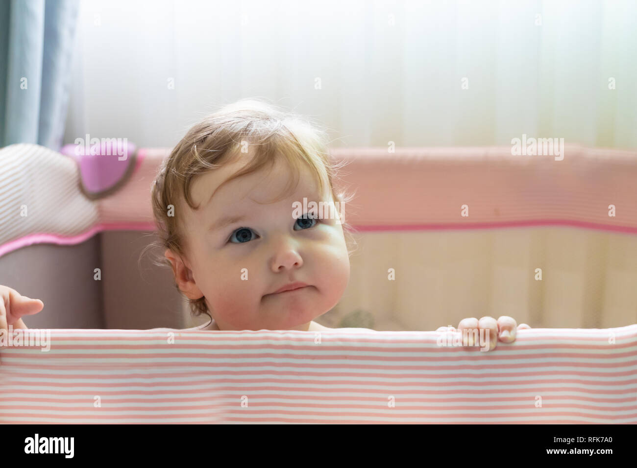 Cute lachendes Baby stehen in einem rosa Bett. Rosa Kinderzimmer für kleine Kinder. Ein kleines Mädchen lernt in ihrer Krippe zu stehen. Ein Baby steht in einer Krippe auf einem sonnigen Schlafzimmer. Das Kind ist traurig und lächelnd, wachte nur nach oben Stockfoto