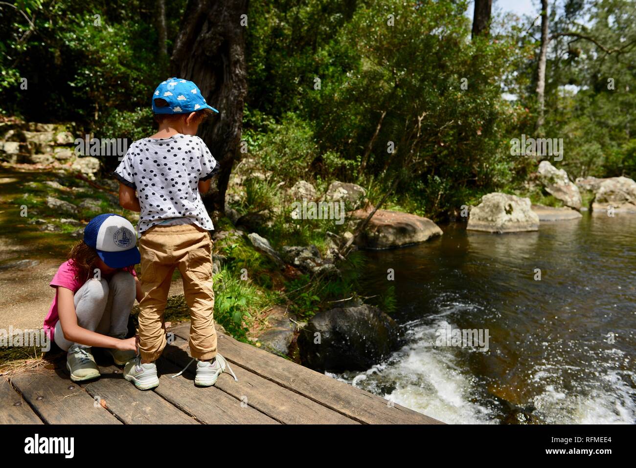 Ein Kind hilft, ein anderes Kind binden Ihre Schnürsenkel auf einem kleinen hölzernen Brücke über einen Fluss, Eungella National Park, Queensland, Australien Stockfoto
