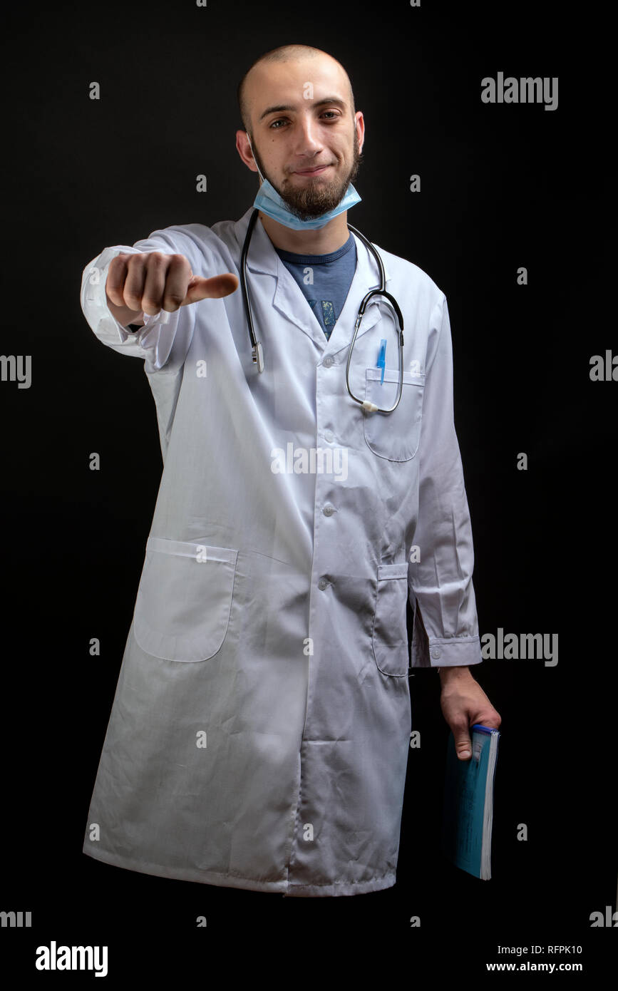 Junge männliche Arzt Holding eine Datei, mit Stethoskop und Maske displaing einen Finger Daumen horizontal. Proffesional healthcare Hintergrund Stockfoto