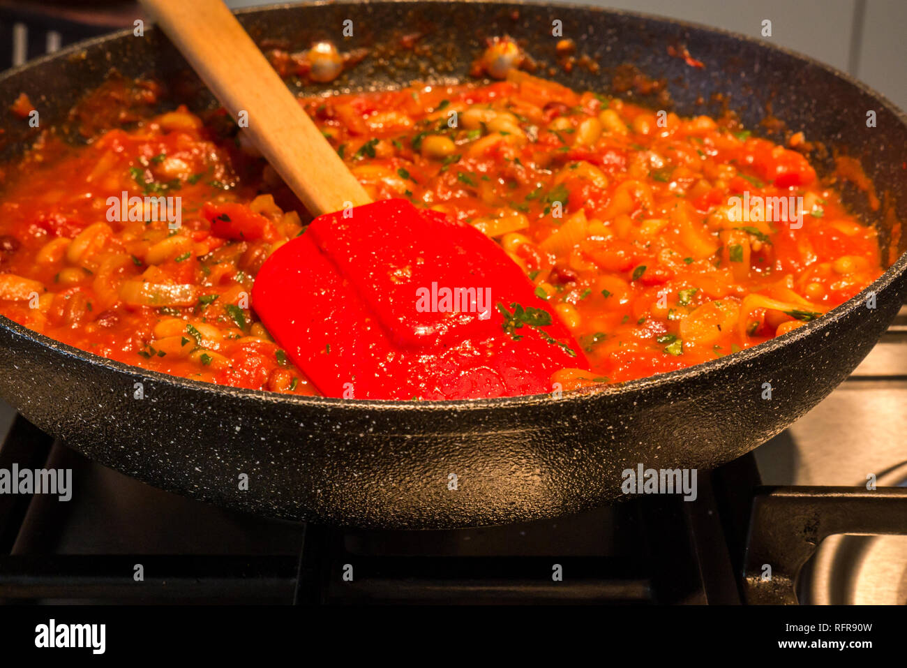Close up Vorbereitung der Italienischen Eintopf mit Tomaten, Bohnen, Kräuter und die Zwiebel Sauce in der Pfanne auf Gas Ring mit Holz behandelt Spachtel Stockfoto