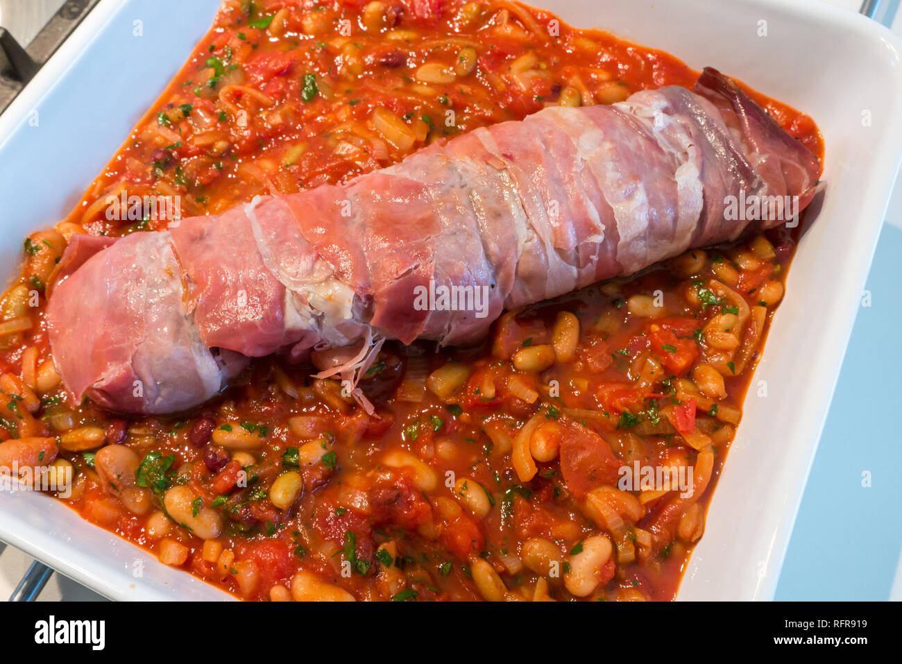 Close up Italienische Gericht aus Schweinefilet in Prosciutto eingewickelt bereit zum Kochen auf Bett von Tomaten, Bohnen, Kräutern und Knoblauch in weißen Keramikschale Stockfoto