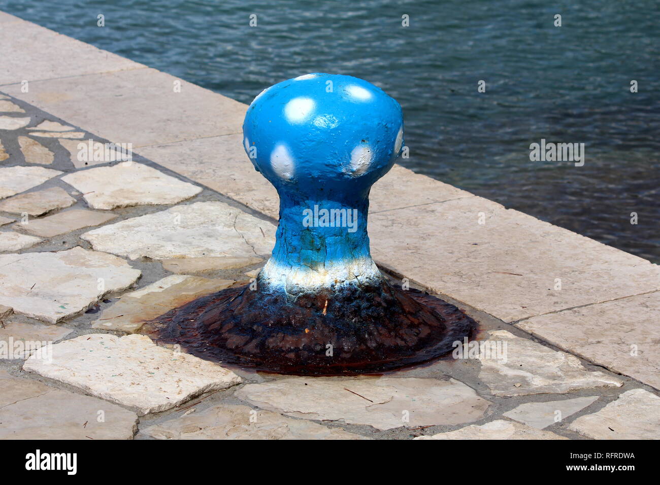 Verrostete alte Bügeleisen bootsanlegeplatz Poller geformt und wie blaue Pilz mit weißen Flecken auf traditionellen Stein Pier montiert lackiert neben Meer an kalten sonnigen Tag Stockfoto
