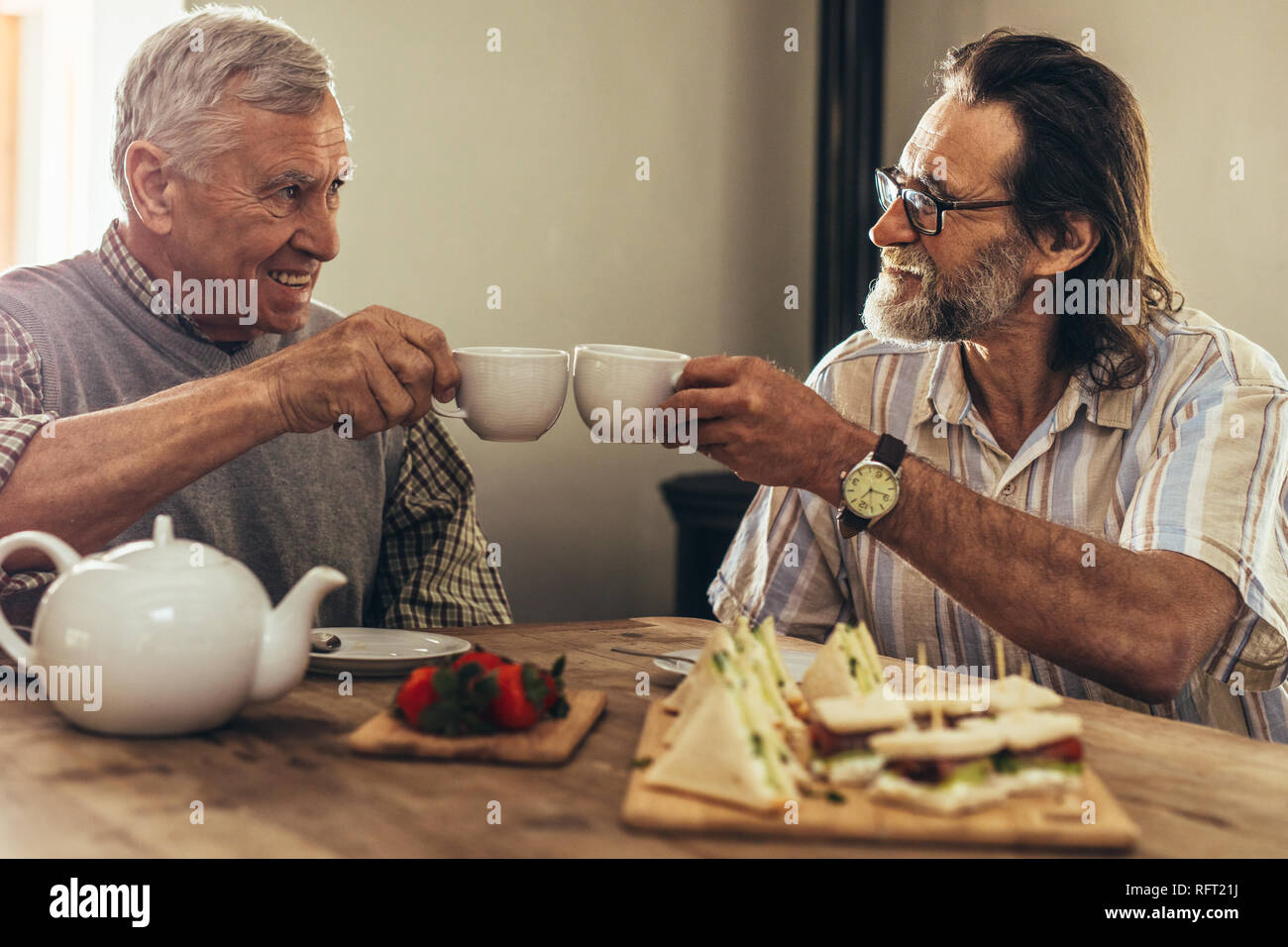 Zwei beste Freunde toasten Kaffee Tassen, während bei Tisch drinnen sitzen. Ältere Männer genießen Sie Kaffee und Snacks zusammen zu Hause. Stockfoto