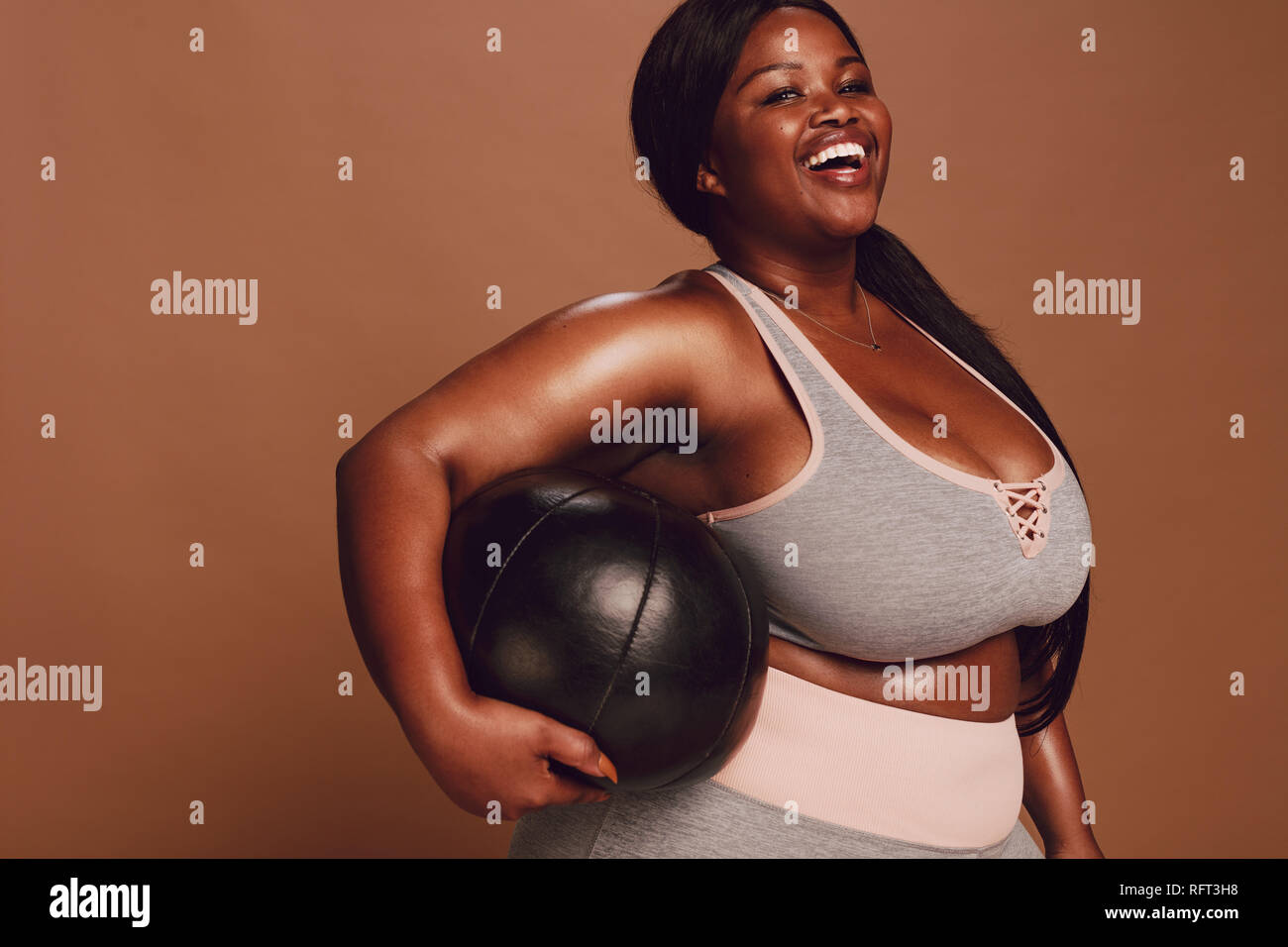 Freundliche große afrikanische Frau mit einem medizinball auf braunem Hintergrund. Übergrößen Frauen im Sport Bekleidung suchen an Kamera und Lachen. Stockfoto