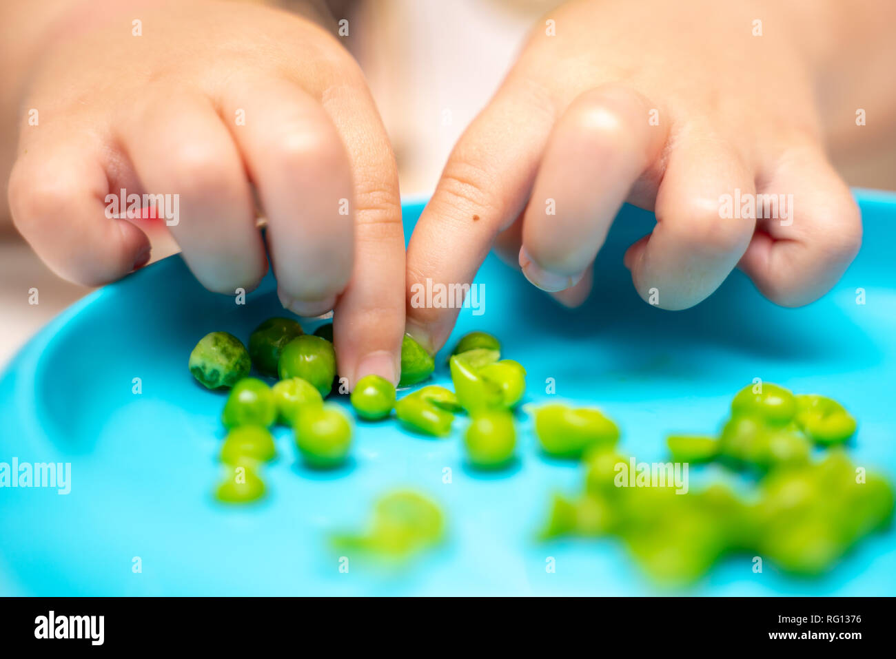 Die Hände des jungen Kindes aufnehmen, spielen und essen Erbsen zum Abendessen wird von einem blauen Platte Stockfoto