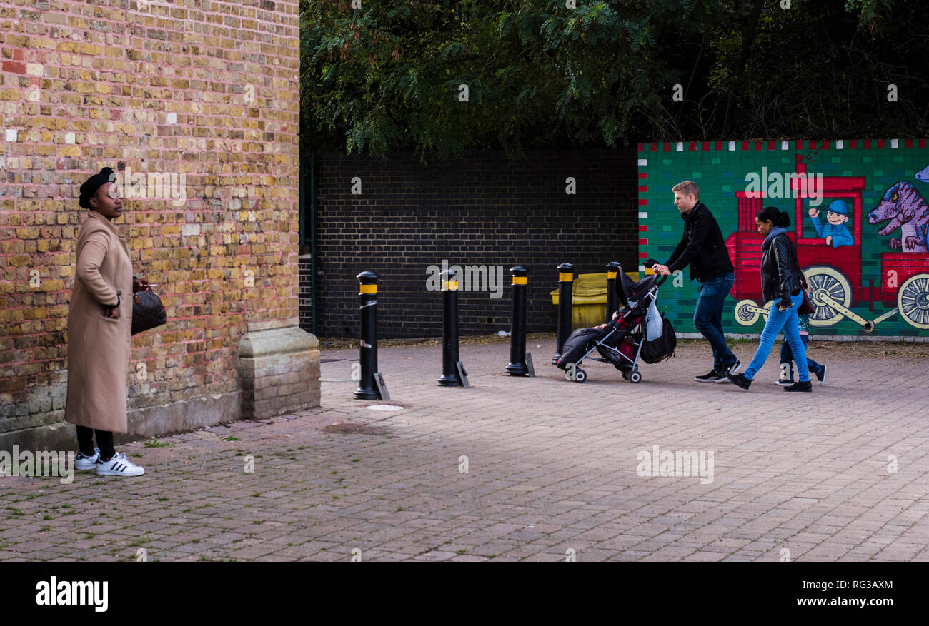 Frau stand neben der Mauer, auf der Suche nach Abstand, junge Familie wandern im Hintergrund, schieben Kinderwagen, Crystal Palace, London, England, Großbritannien Stockfoto