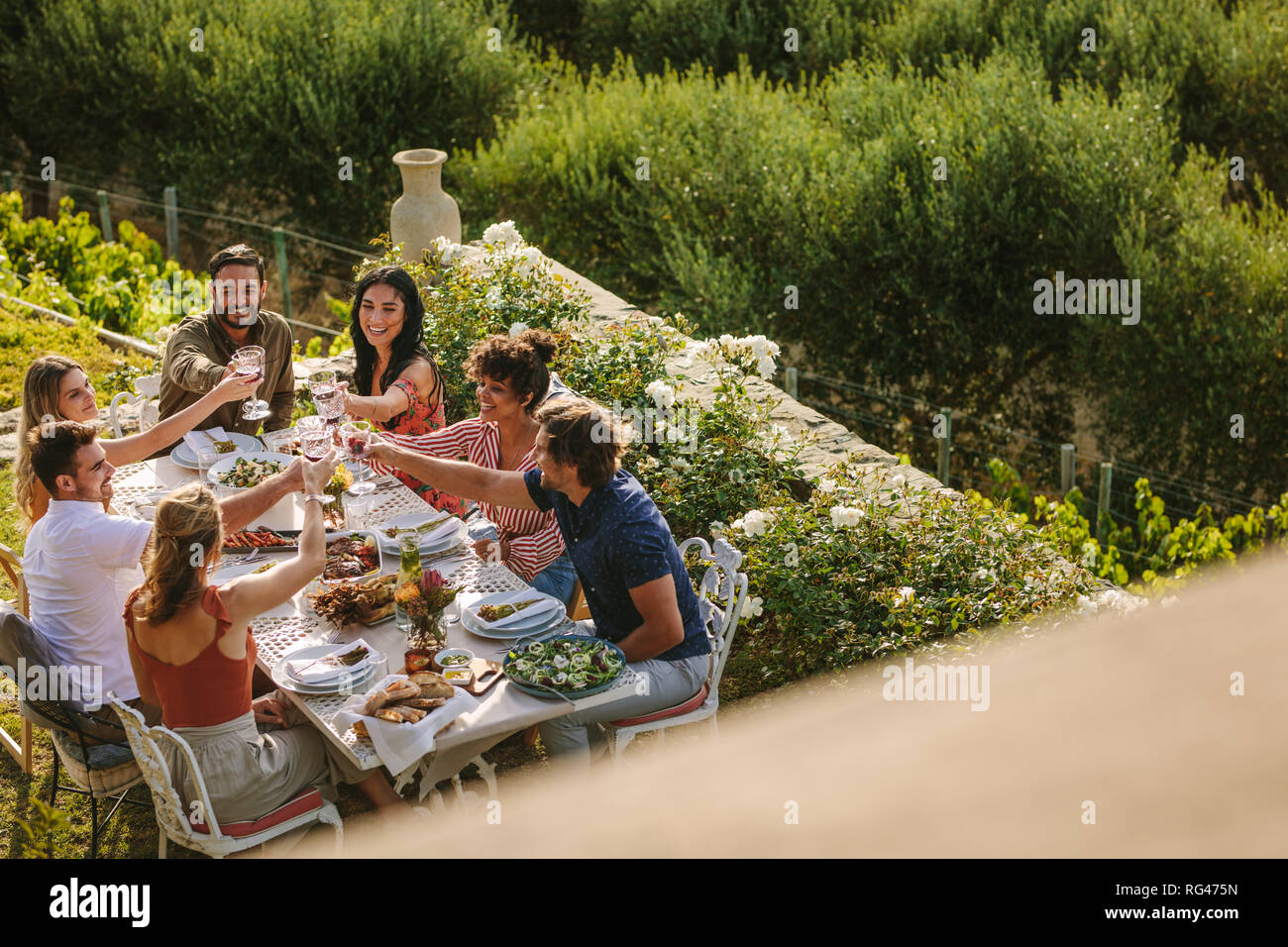 Hohe Betrachtungswinkel der jungen Menschen jubeln mit Getränken bei einem Abendessen im Freien. Gruppe Männer und Frauen sitzen am Tisch und toasten Wein Glas Stockfoto