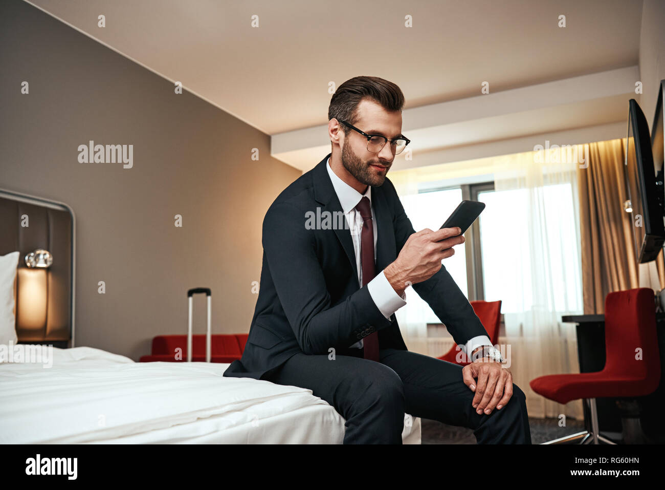 Entspannen Sie sich nach langen Geschäftsreise. Geschäftsmann in Anzug sitzen mit Smartphone auf dem Bett im Hotel Zimmer und chatten mit einem Freund Stockfoto