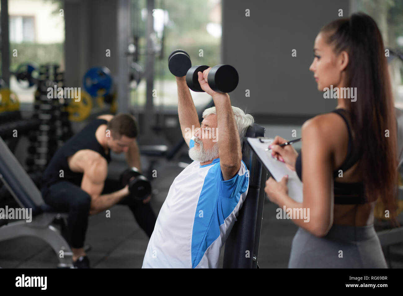 Horisontal Detailansicht von drei Leuten im Fitnesscenter zu trainieren. Athletische junge Frau stehen und weg schauen mit Tablette, während Ihr Client elderler Mann tun, ausüben. Konzept des Lebensstils. Stockfoto