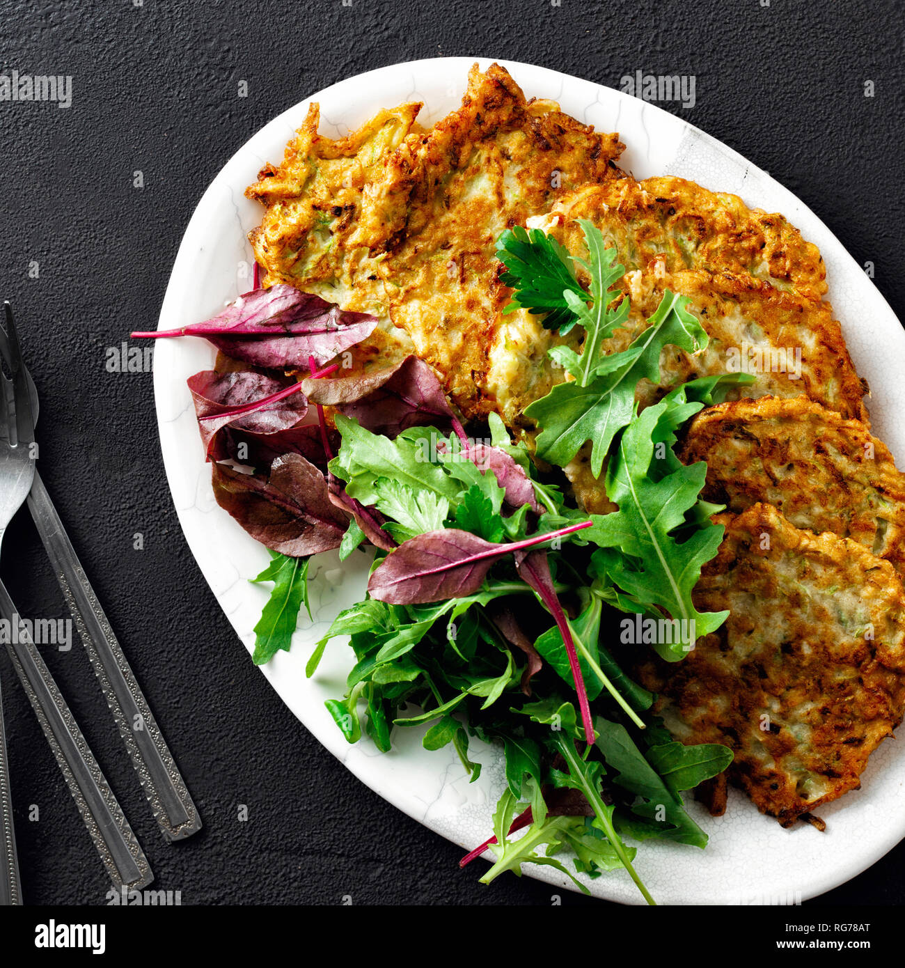 Gebratene zucchini Pfannkuchen serviert auf Platte mit Salat auf dunklem Hintergrund der Ansicht von oben. Comfort food Konzept Stockfoto