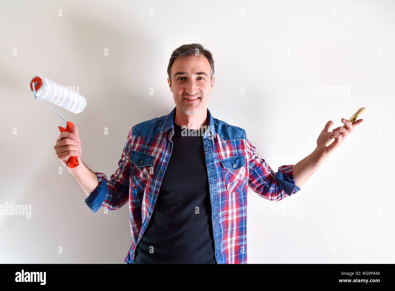 Lächelnder Mann mit Rolle und Pinsel und weiße Wand hinter sich. Diy home Konzept. Vorderansicht. Horizontale Komposition. Stockfoto