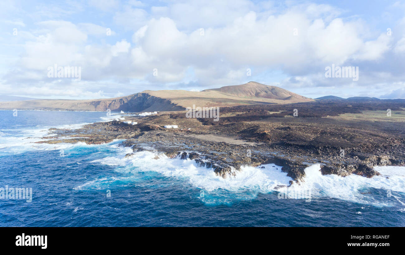 Luftaufnahme von einem Fischerdorf auf einem felsigen Ufer durch hohe Wellen geschlagen, mit vulkanischen Berge am Horizont,, Lanzarote, Kanarische Inseln, Spanien. Stockfoto