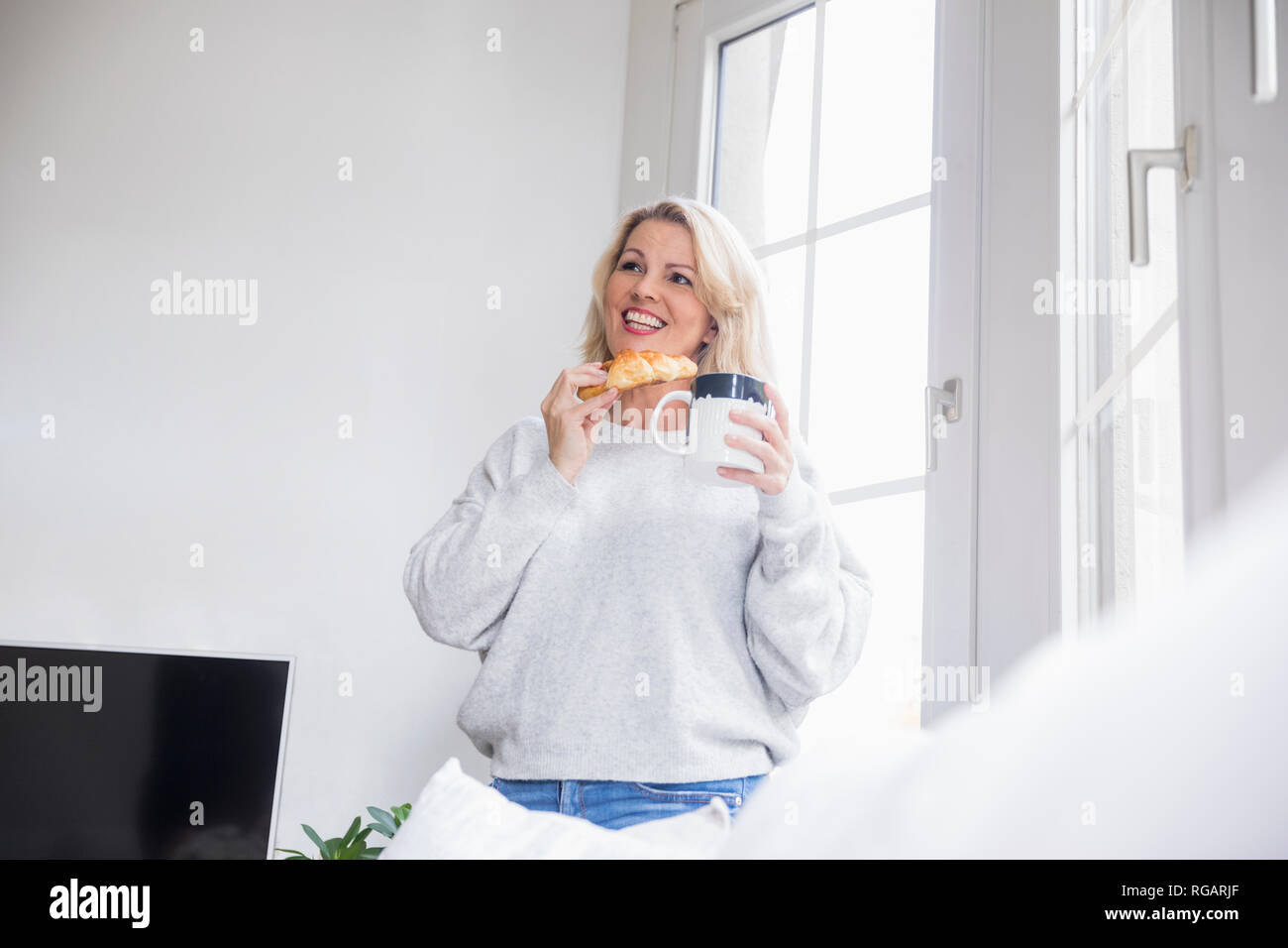 Portrait von lächelnden Blonde reife Frau mit Croissant und Kaffee Tasse zu Hause Stockfoto