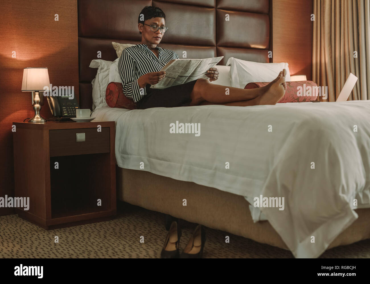 Reife Geschäftsfrau Entspannung im Bett und liest Zeitung in modernen Hotelzimmer. Afrikanische Frauen in formelle Kleidung Entspannung auf Bett mit Zeitung. Stockfoto