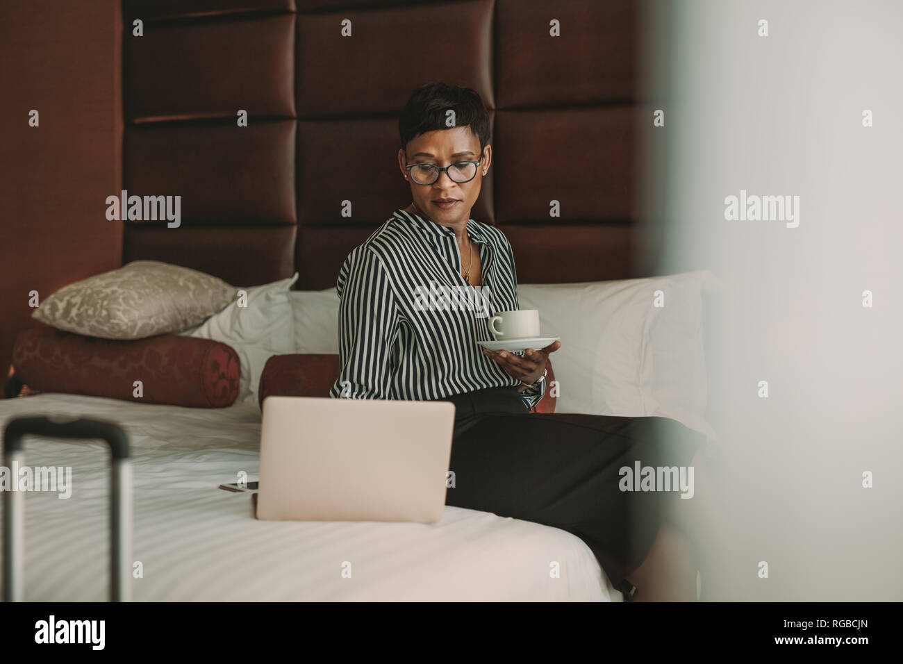 Afrikanische Frau in formelle Kleidung sitzen auf dem Bett mit Kaffee im Hotel Zimmer mit Laptop. Geschäftsfrau arbeiten vom Hotelzimmer. Stockfoto