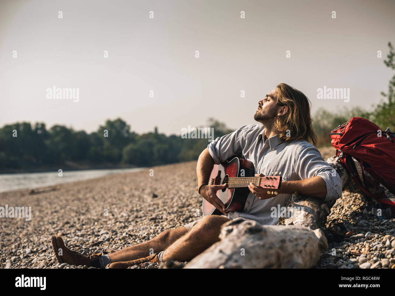 Junger Mann am Flußufer Gitarre spielen Sitzen Stockfoto
