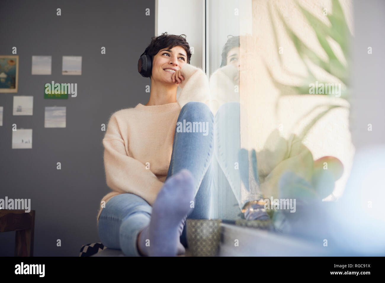 Lächelnde Frau zu Hause mit Kopfhörern am Fenster sitzen Stockfoto