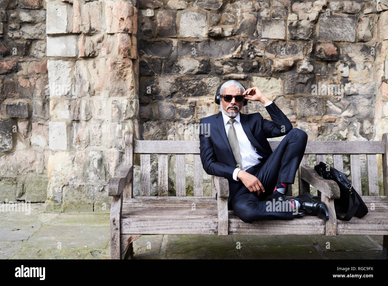Portrait von gut gekleideten älteren Geschäftsmann mit Sonnenbrille und Kopfhörer sitzt auf der Bank Stockfoto