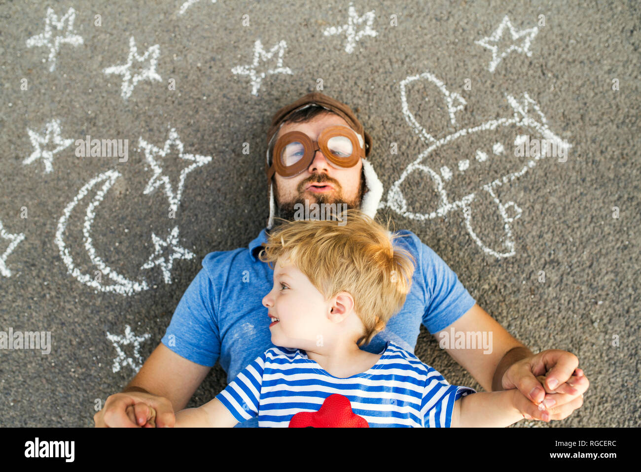 Portrait von reifer Mann, der Pilot hat und seinem kleinen Sohn liegend auf Asphalt mit Flugzeug, Mond und Sternen bemalt Stockfoto