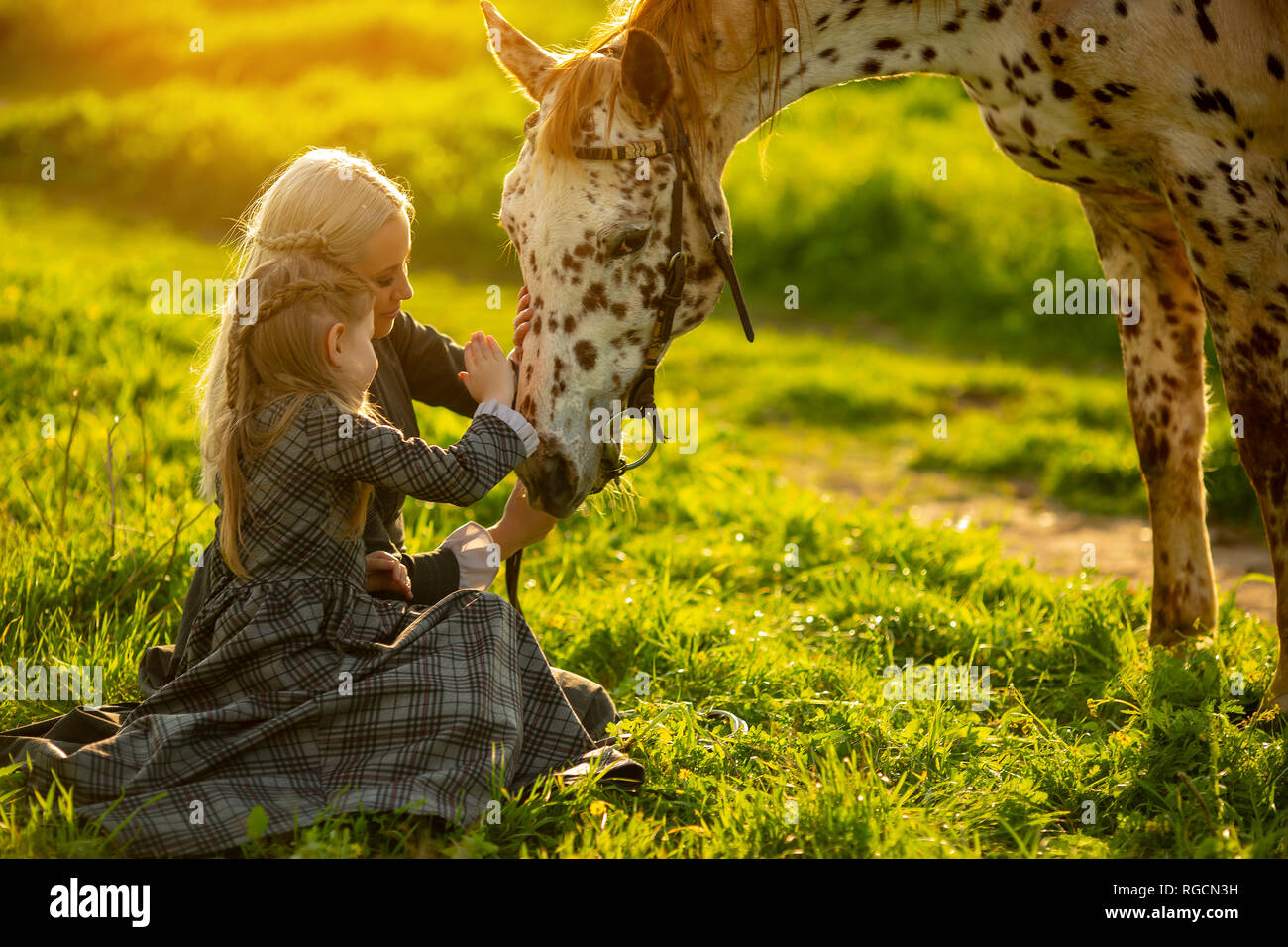Seitliche Sicht auf eine junge Mutter mit einem kleinen Mädchen in den Kleidern Schlaganfall ein beschmutztes Pferd auf einer grünen Wiese Stockfoto