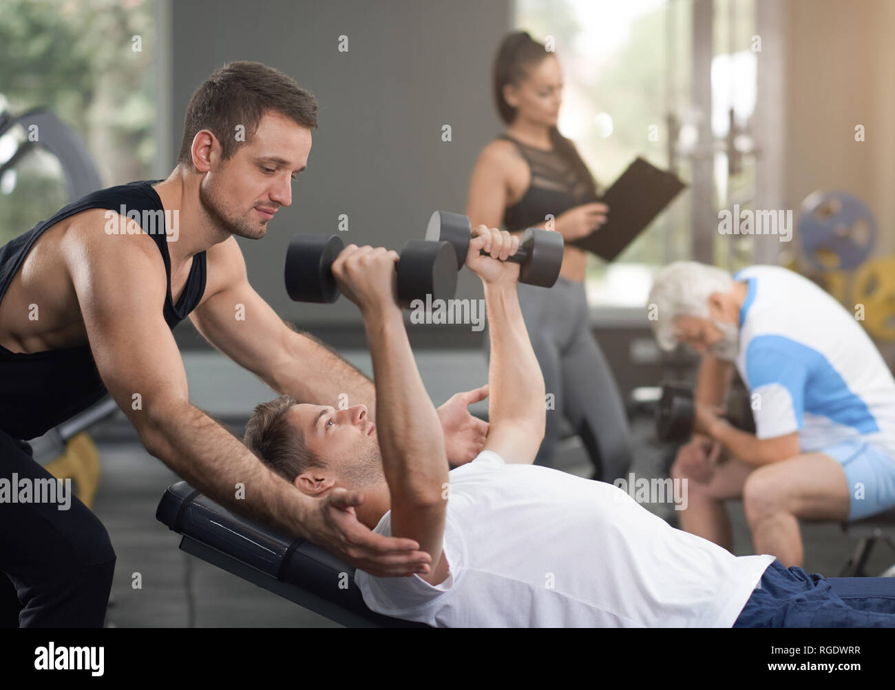 Seitenansicht der Mann im weißen T-Shirt liegen auf der Werkbank, holding hands up mit Hanteln. Muskulöse Trainer helfen Client mit sportlicher Ausstattung. Konzept der modernen Fitnessraum und Sport. Stockfoto