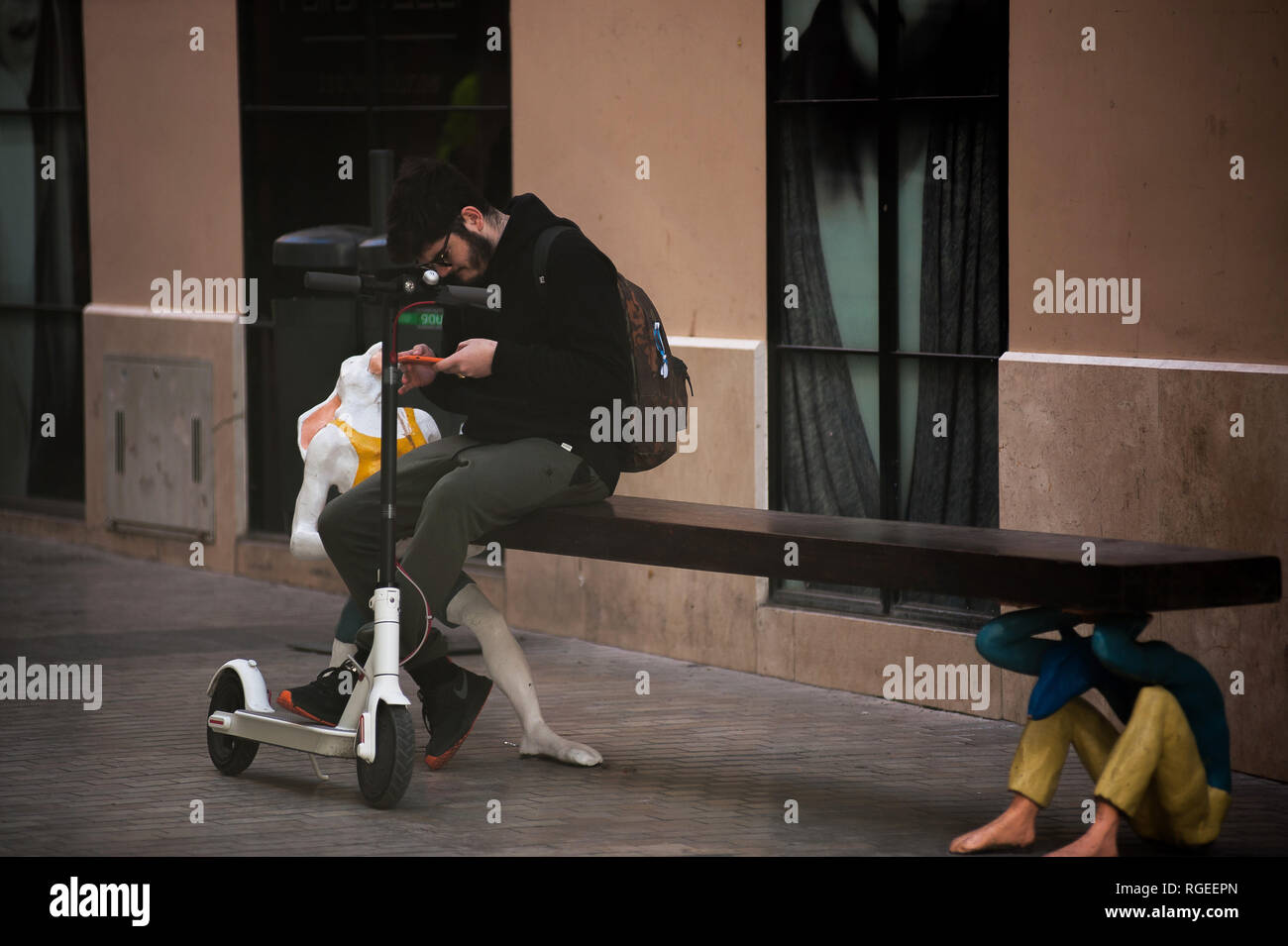 Ein Mann ruht und Kontrolle sein Handy auf einer Bank mit einem kostenlosen Elektroroller ist auf der Straße gesehen. Der spanische General Traffic Management wird von nun an einen Führerschein für die elektroroller und eine Versicherung unter anderem Maßnahmen bedarf, entsprechend einer neuen Dekret von der spanischen Regierung. In den letzten Monaten hat mehr Unfälle mit Elektroroller passiert, und ein Tod im vergangenen Jahr. Die Regierung erwarten, wie dies der Verwendung dieser Transporte zu regulieren und die Sicherheit verbessern. Stockfoto