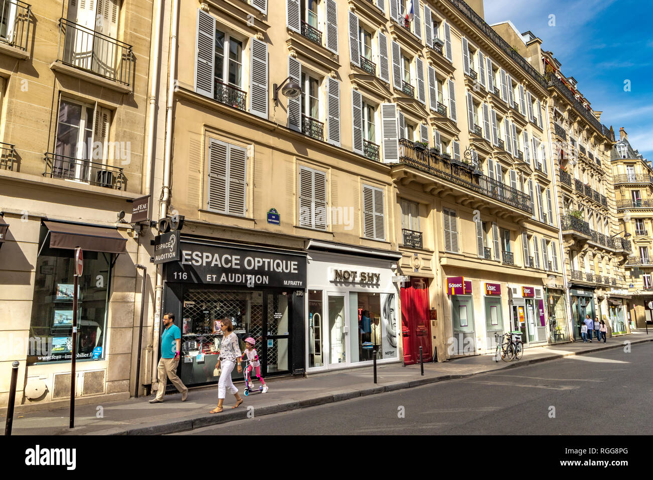 Ein Mann und eine Frau mit einem kleinen Kind auf einem Roller vorbei gehen die Geschäfte und Wohnhäuser auf der Rue Saint-Dominique, Paris, Frankreich Stockfoto