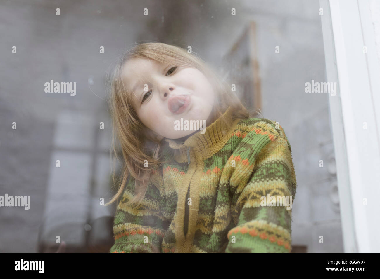 Portrait von kleinen Mädchen Zunge heraus haften, während sie aus dem Fenster Stockfoto