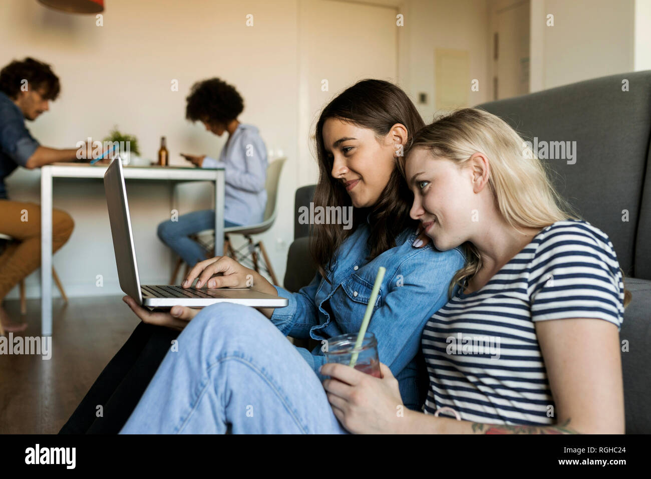 Zwei lächelnde junge Frauen sitzen auf dem Boden teilen Laptop mit Freunden im Hintergrund Stockfoto