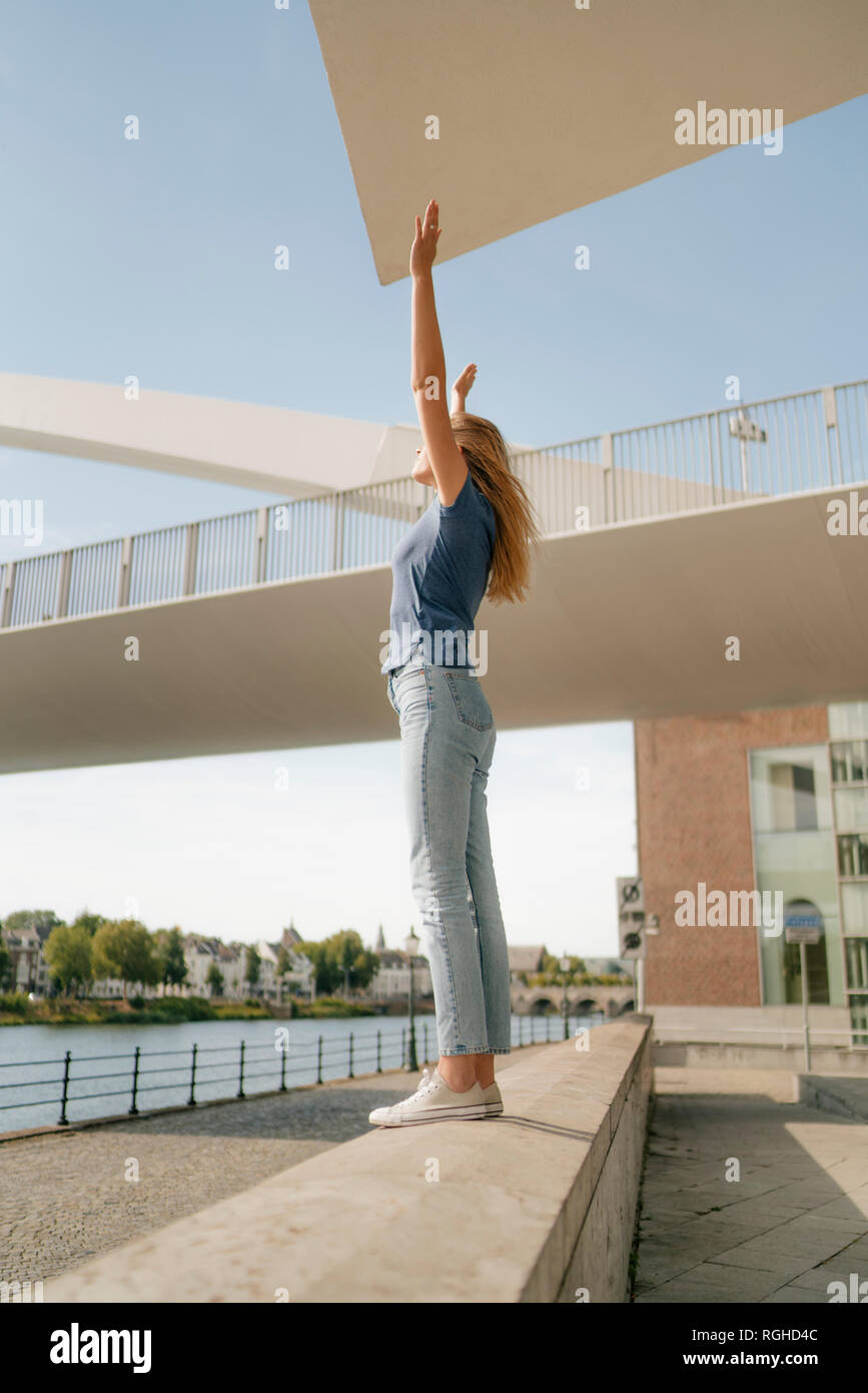 Niederlande, Maastricht, junge Frau, die an der Wand am Flußufer mit erhobenen Armen Stockfoto