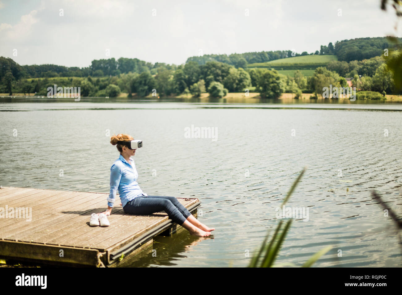 Frau mit VR-Brille sitzen auf Steg an einem See mit den Füßen im Wasser Stockfoto