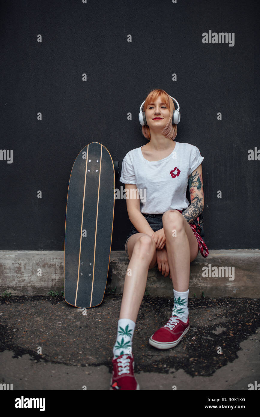 Lächelnde junge Frau mit Skateboard und Kopfhörer sitzen unten Hören von Musik Stockfoto