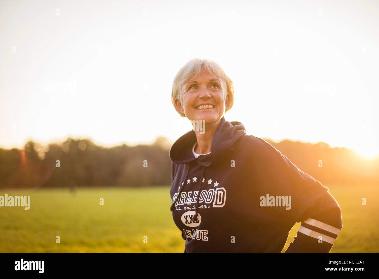 Lächelnd senior Frau trägt ein Hoodie stehend auf ländlichen Wiese bei Sonnenuntergang Stockfoto