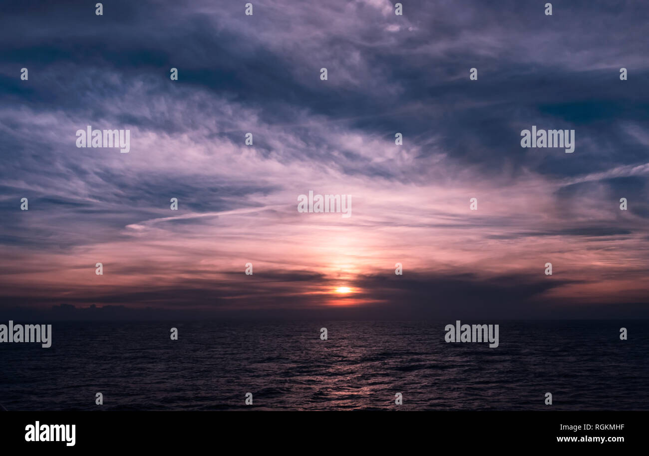 Unglaubliche Sonnenuntergang Himmel mit Wolken und den Farben lila und rot gefüllt. Kreuzfahrtschiff Blick auf einen wunderschönen Abendhimmel über der Meeresoberfläche widerspiegeln. Stockfoto
