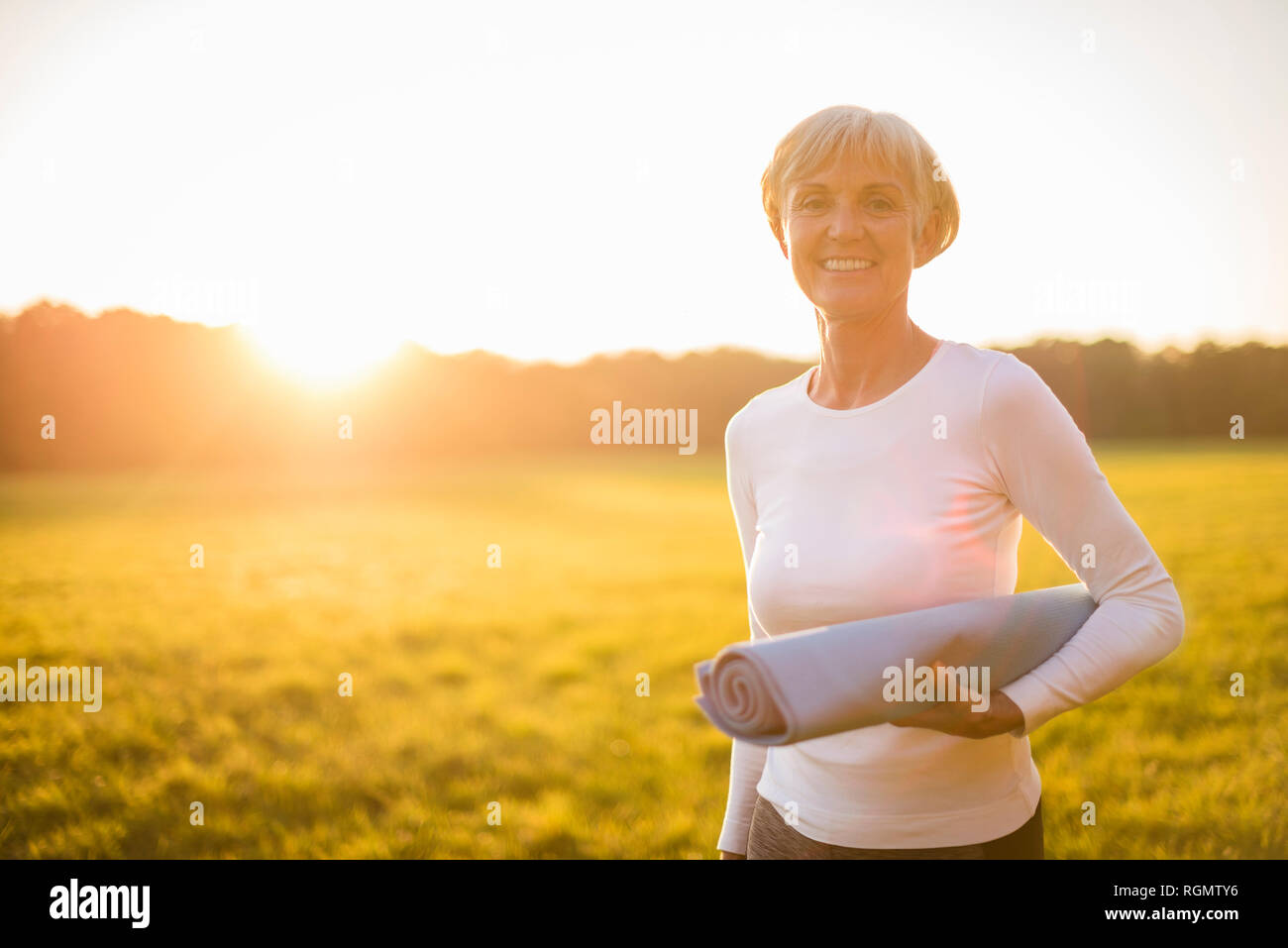 Portrait von lächelnden älteren Frau mit Yoga Matte auf ländlichen Wiese bei Sonnenuntergang Stockfoto