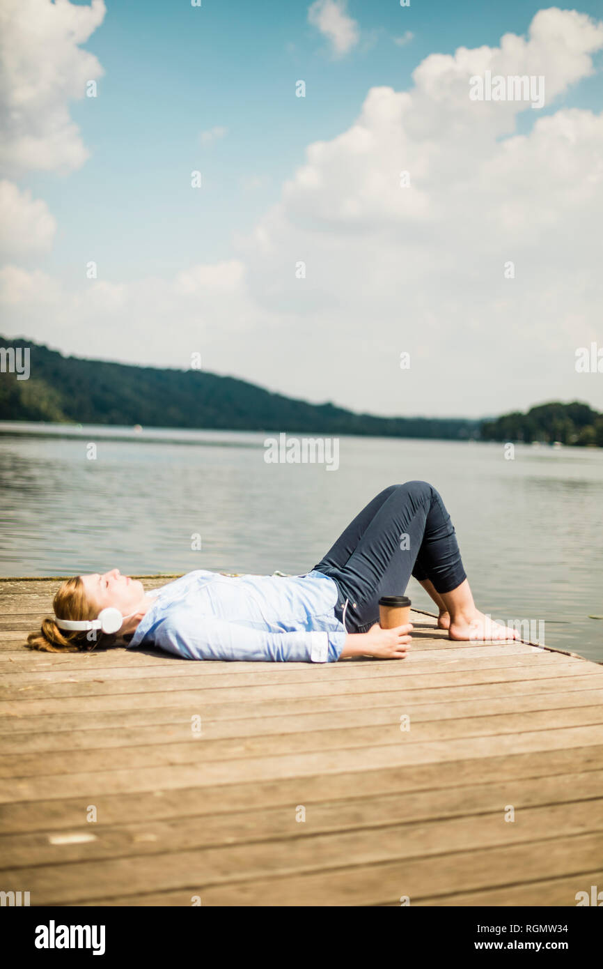 Liegende Frau auf Steg an einem See mit Kopfhörern und Kaffee zum Mitnehmen Stockfoto
