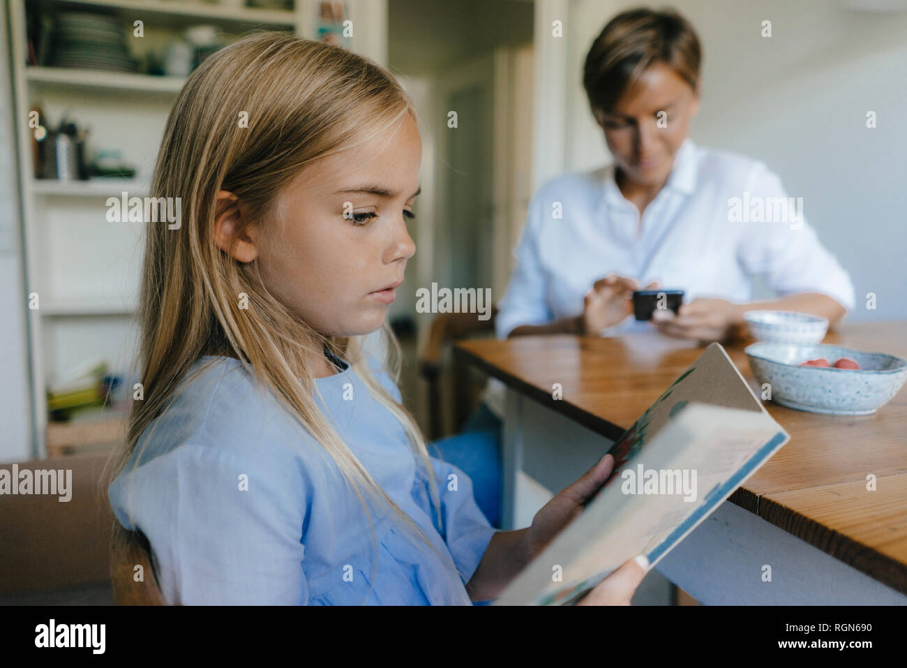Mädchen mit Buch am Tisch zu Hause sitzen mit Mutter mit Handy im Hintergrund Stockfoto