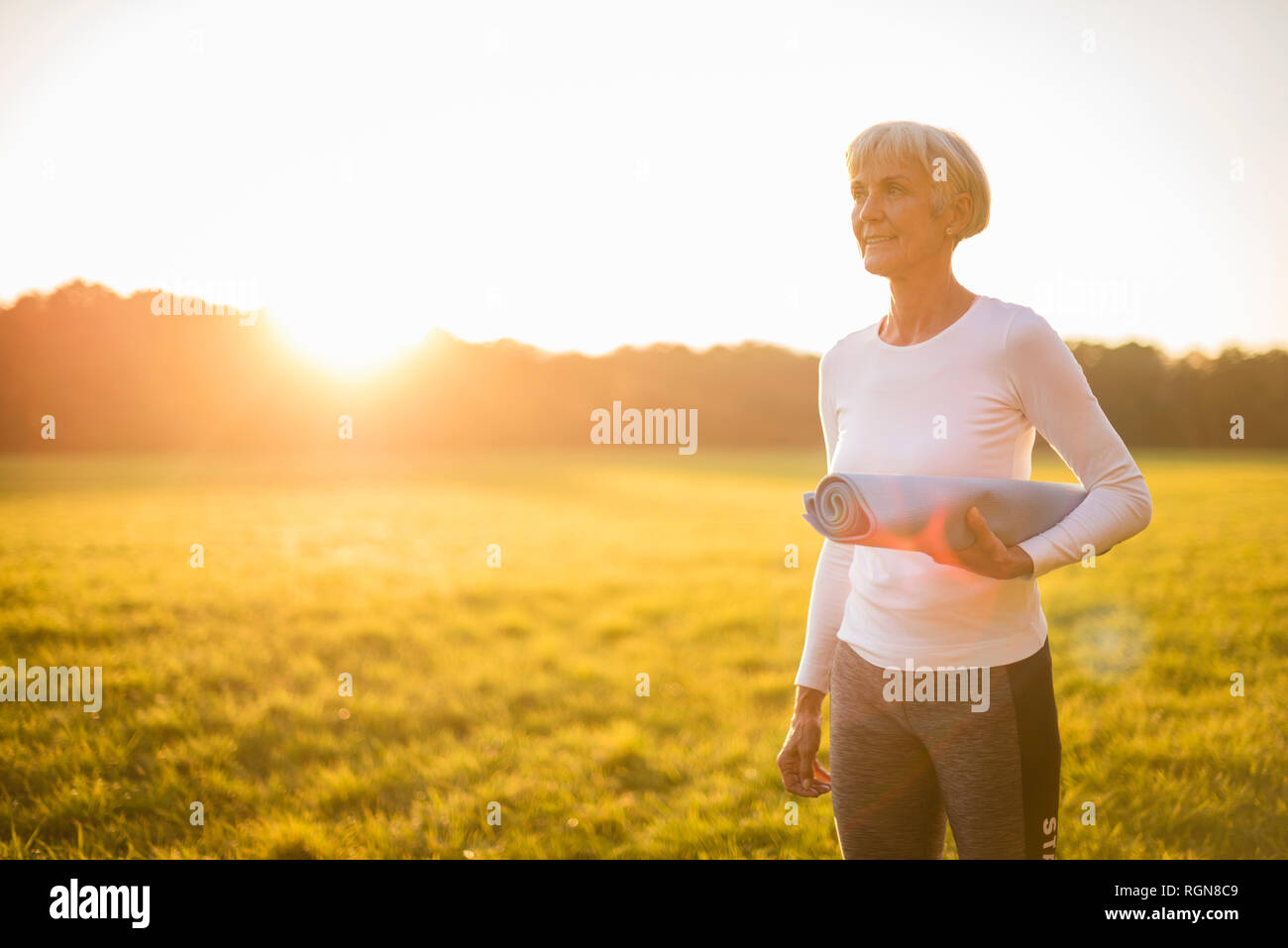 Ältere Frau mit Yoga Matte auf ländlichen Wiese bei Sonnenuntergang Stockfoto